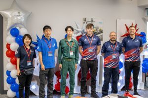 Group of six people standing in front of a balloon decoration, three in matching blue uniforms with red and white accents, one in green coveralls, another in casual attire, and one in a polo shirt.